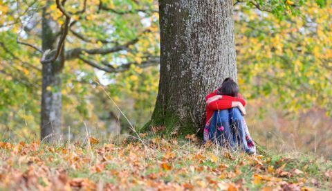 Una persona sentada bajo un árbol en otoño con la cabeza apoyada entre las piernas. Shutterstock / Juice Flair